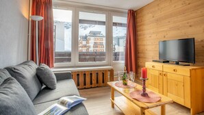 A cozy living room with a grey sofa, a wooden TV stand, a window with a view of buildings, and a table with glasses and a candle.
