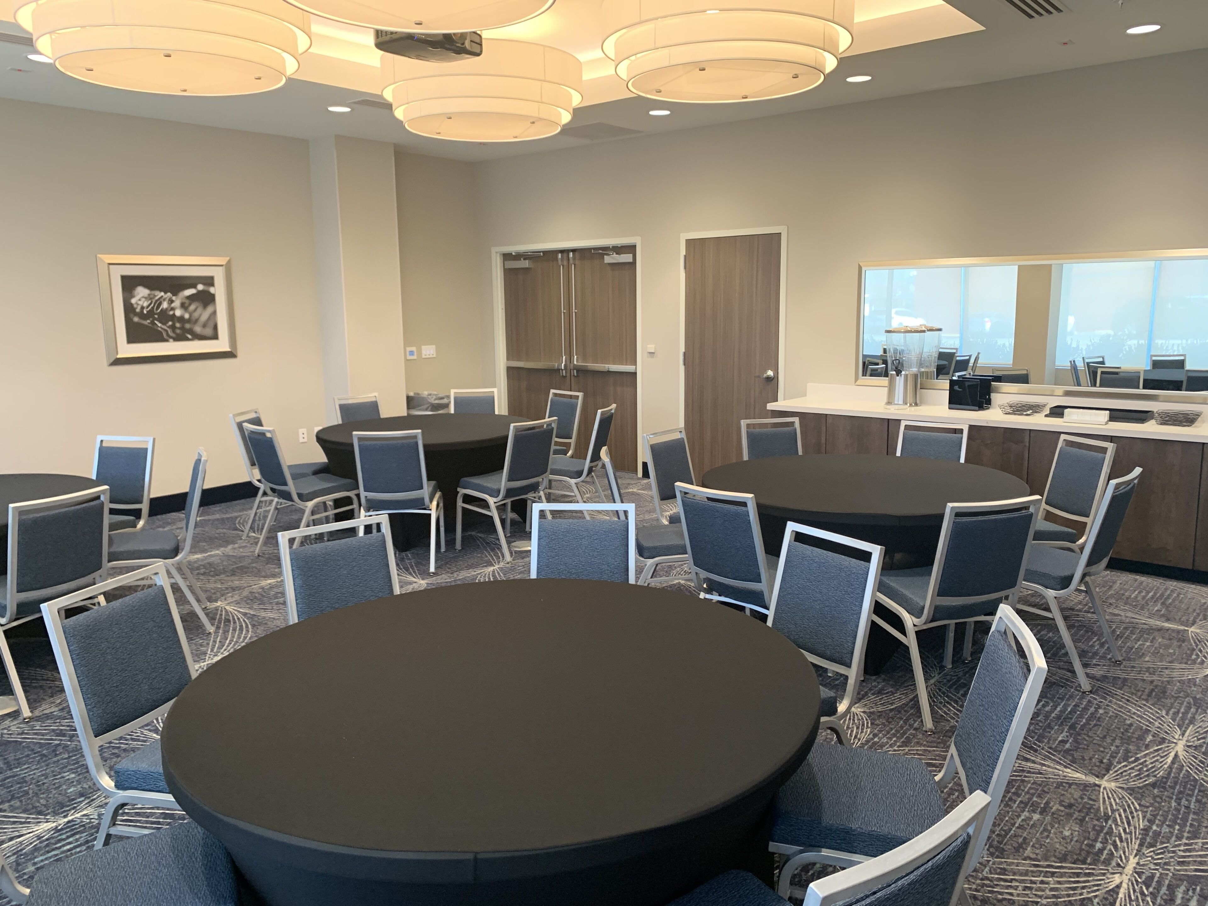 A conference room with a large round table surrounded by chairs, a kitchenette area, and a large window providing natural light.
