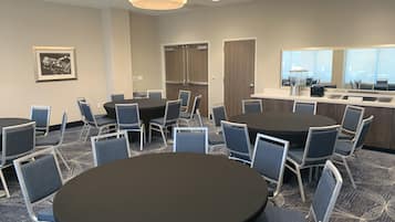 A conference room with a large round table surrounded by chairs, a kitchenette area, and a large window providing natural light.