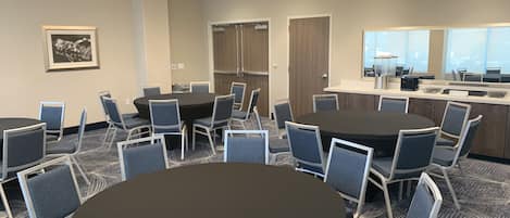 Un salón de conferencias con una amplia mesa redonda rodeada de sillas, una zona de kitchenette y un gran ventanal que proporciona luz natural.