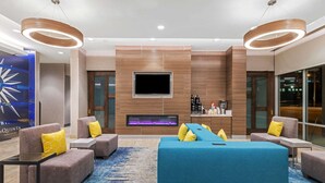 A modern hotel lobby with a large blue sofa, two armchairs, a flat-screen TV, a fireplace, and a coffee station.