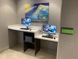 Un espacio de trabajo con dos escritorios, cada uno con computadora, teclado y ratón. Hay una impresora debajo de uno de los escritorios. Se ve un letrero en la pared que dice 'BUSINESS CENTER'.