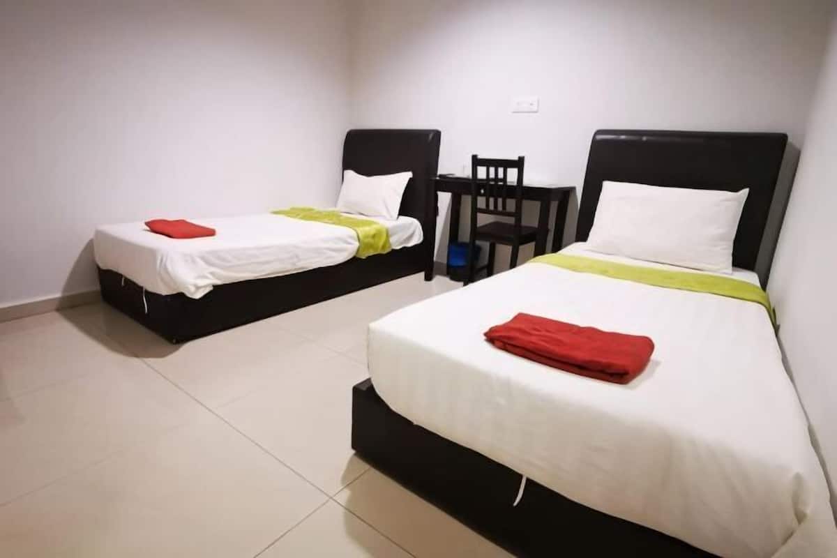Duas camas de solteiro com cabeceiras pretas e roupa de cama branca, uma mesinha e uma cadeira num quarto com paredes brancas e chão de azulejos.