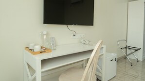 Une chambre d’hôtel avec un bureau blanc, un téléviseur à écran plat fixé au mur, un téléphone, une petite table avec un plateau contenant des tasses et un verre, et une chaise dotée d’un porte-serviettes.