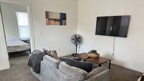 Ein Wohnzimmer mit grauem Sofa, einem kleinen Tisch mit Korb, einer Stehlampe und einem an der Wand befestigten Fernseher.