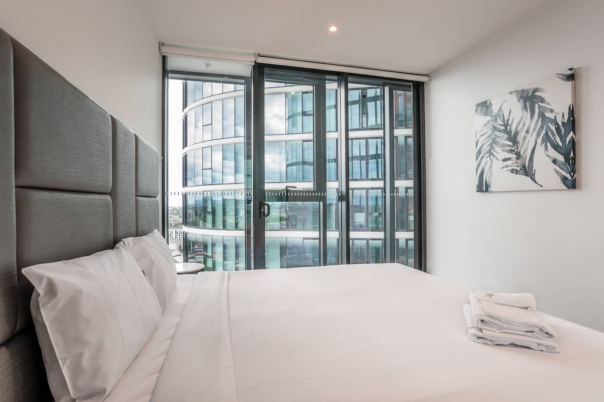 Ein modernes Hotelzimmer mit einem großen Bett, Blick auf ein Gebäude durch raumhohe Fenster und einem Wandbild im tropischen Stil.