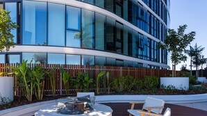 A modern building with large windows and a landscaped outdoor area with a circular seating area and wooden chairs. - FV By Serain Residences (Fortitude Valley)