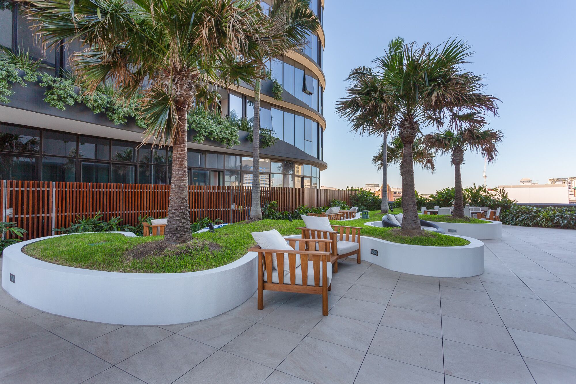 Un espacio al aire libre con sillas de madera y macetas, rodeado por un edificio moderno con ventanas amplias y vegetación.