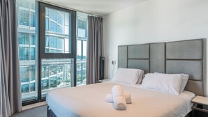 A modern hotel room with a large bed, grey headboard, and a view of a cityscape through floor-to-ceiling windows. - FV By Serain Residences (Fortitude Valley)