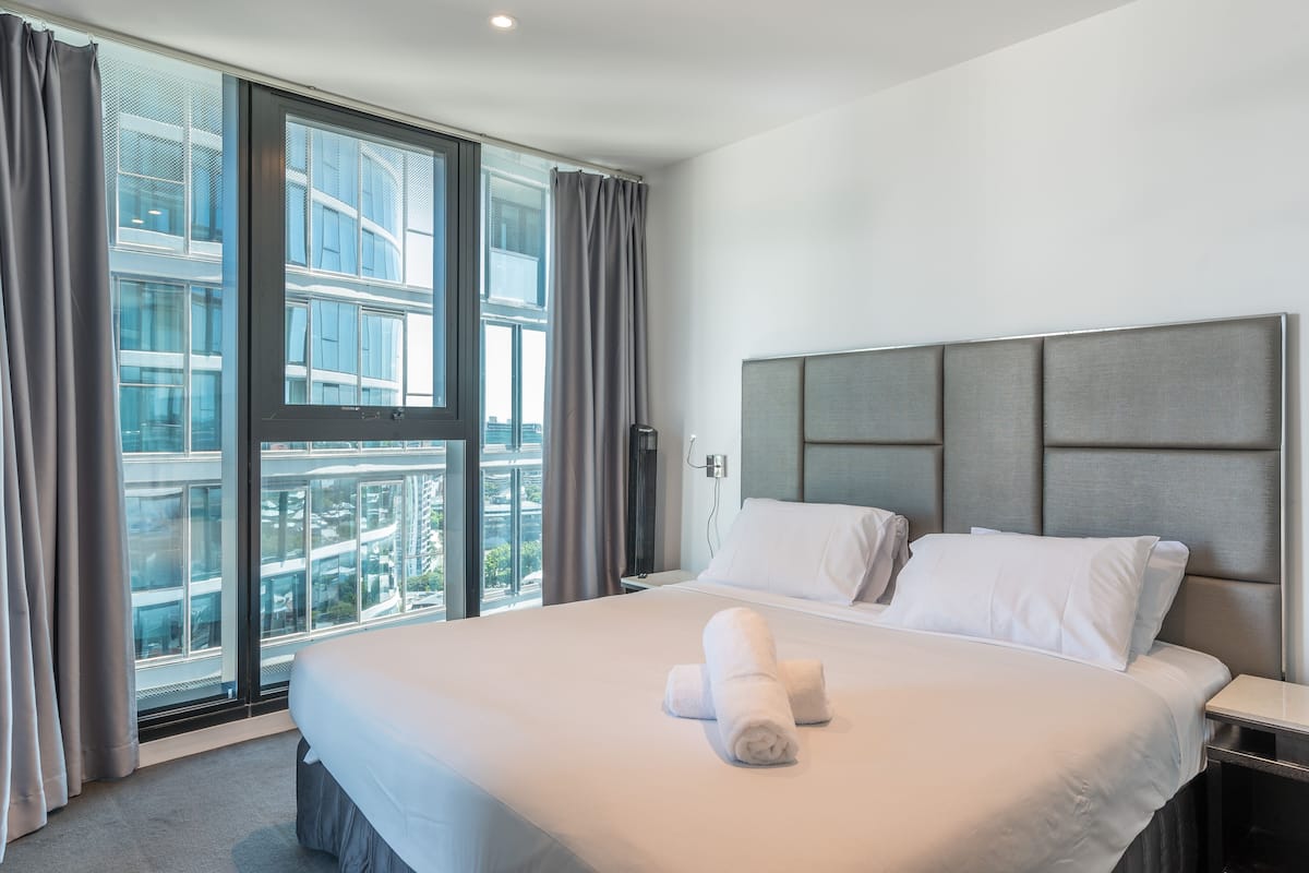 Ein modernes Hotelzimmer mit einem großen Bett, einem grauen Kopfteil und Blick auf eine Stadtkulisse durch raumhohe Fenster.