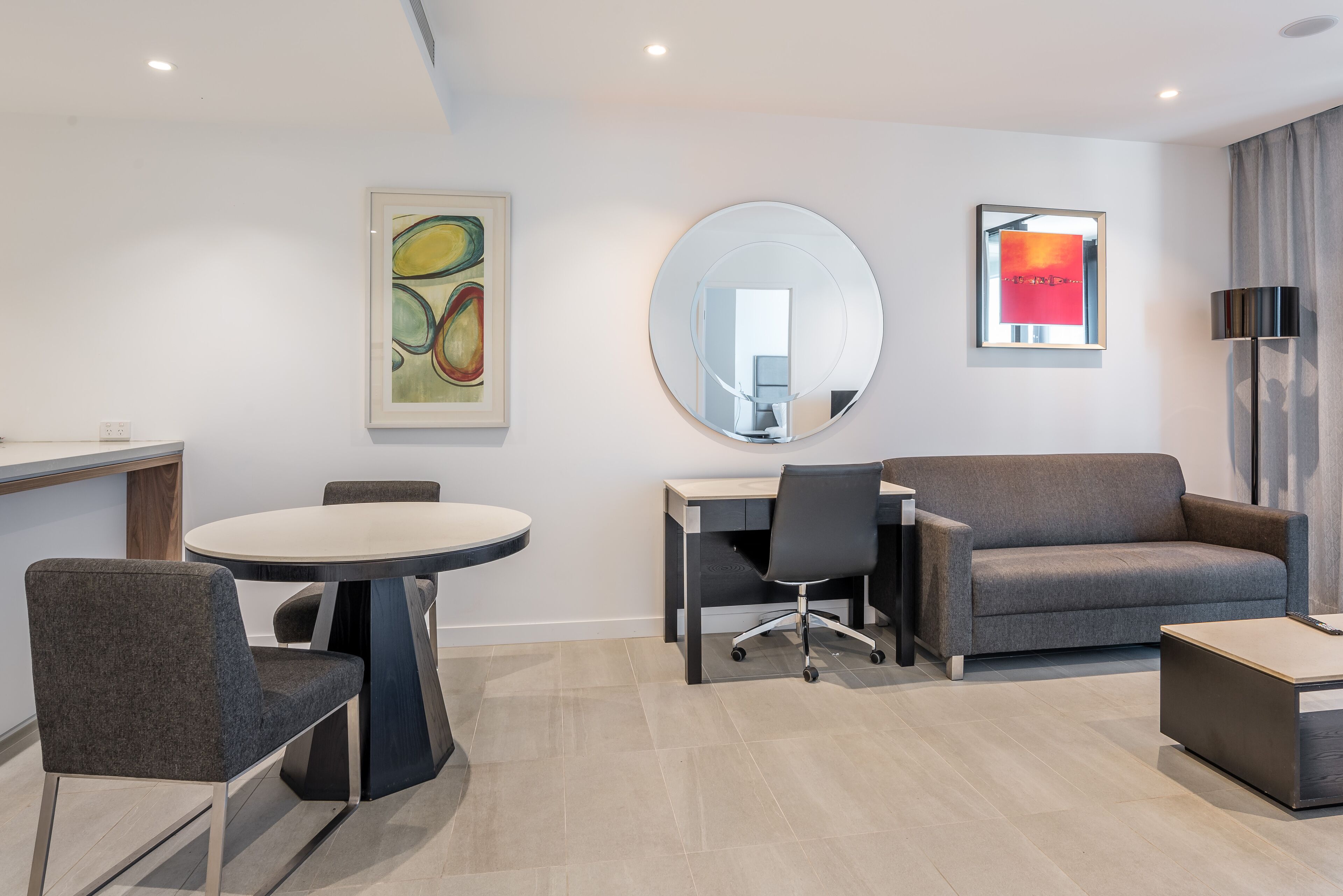 A modern hotel room with a round dining table, a gray sofa, a desk, and a large mirror.