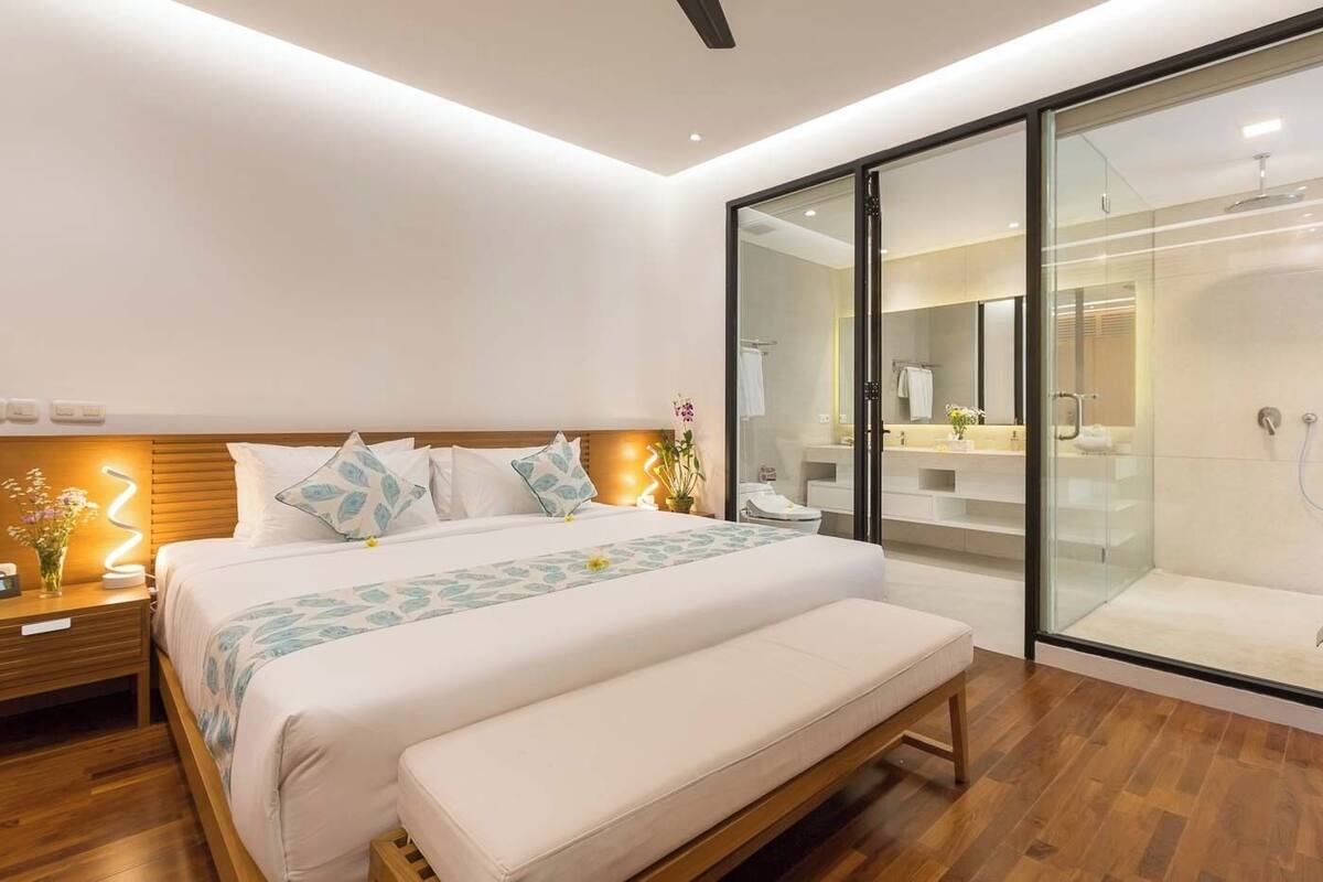 A modern bedroom with a large bed, bedside tables, a bench, a glass-enclosed shower, and a visible bathroom.