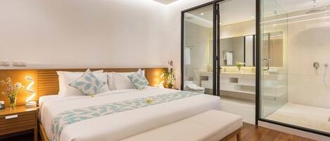 A modern bedroom with a large bed, bedside tables, a bench, a glass-enclosed shower, and a visible bathroom.