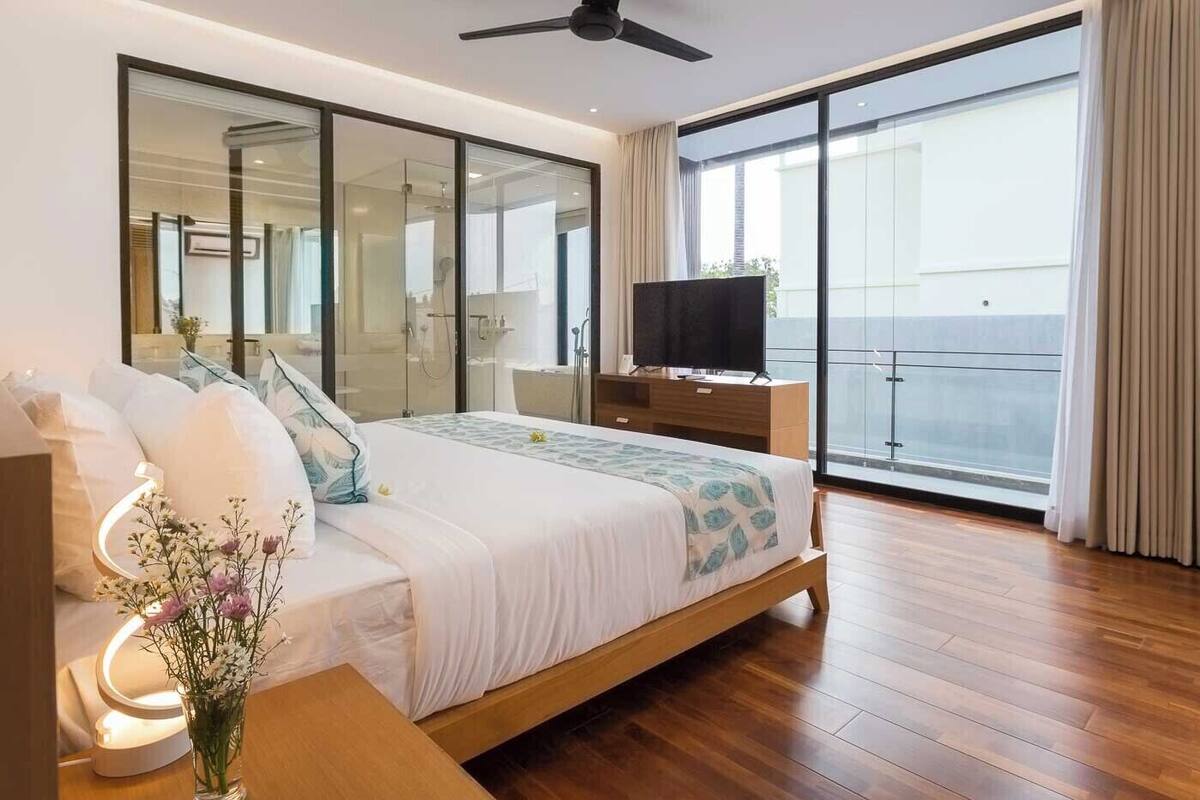 A modern hotel room with a large bed, a TV on a wooden dresser, a glass shower area, and a balcony with a view.