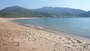 Ein sandiger Strand mit Fußspuren, die zum Wasser führen, umgeben von grünen Hügeln und Bergen.