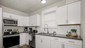 A modern kitchen with white cabinets, stainless steel appliances, a sink, and a window with blinds. - Charming Home | Near Spokane City Center (Spokane)