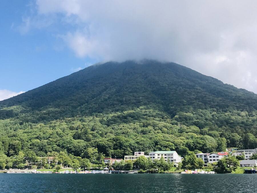 Montanha com base arborizada e edifícios, com vista para um corpo de água.