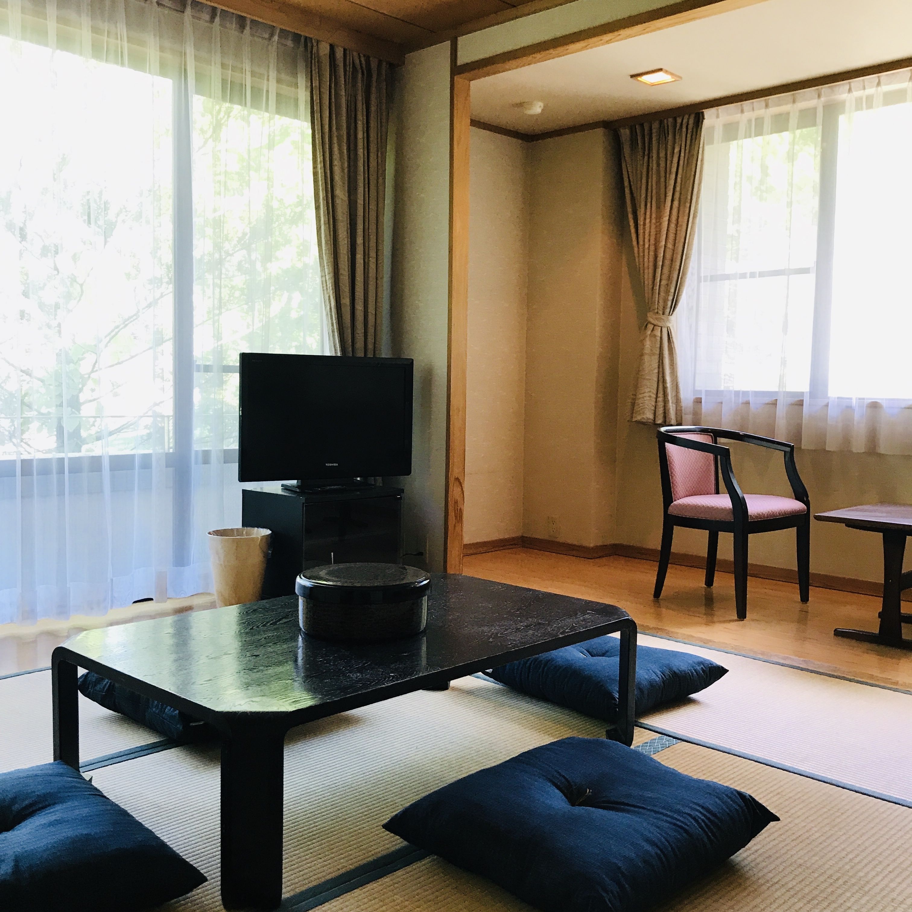 A room with tatami flooring, a television on a stand, a table with cushions, a chair, and a small table.
