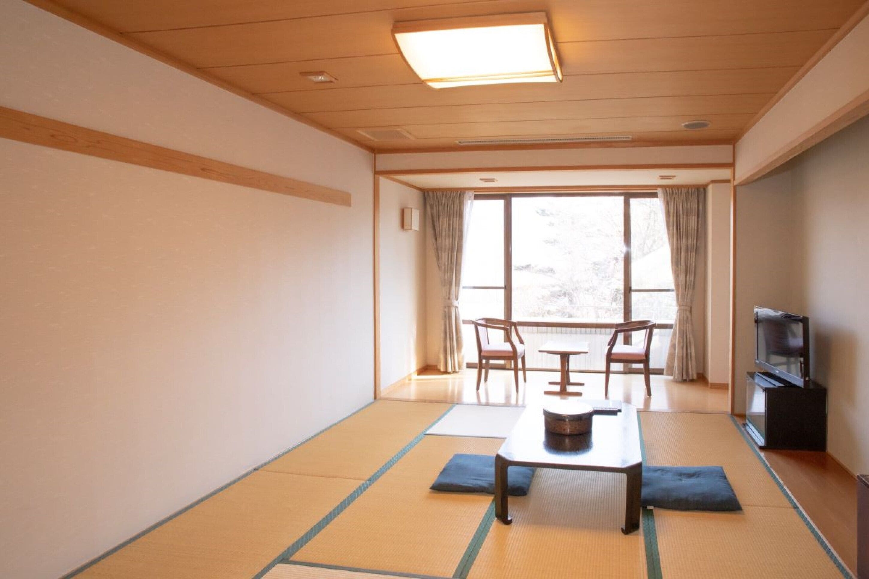 A room with tatami mats, a low table, chairs, a TV, and a large window with curtains.