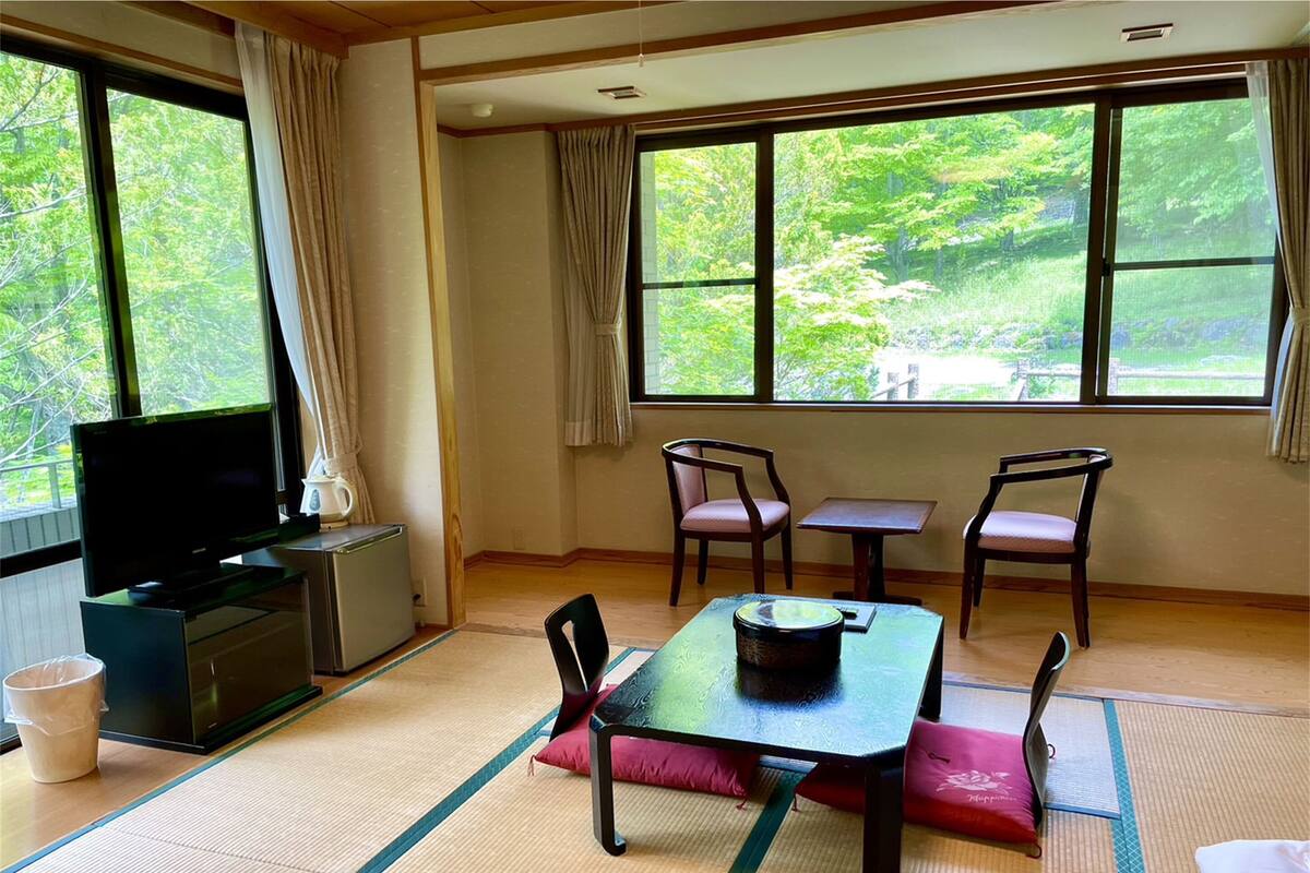 A room with tatami mat flooring, a television on a cabinet, a table with chairs, and large windows with a view of greenery.