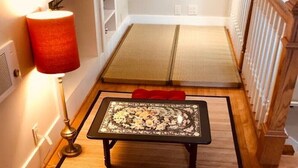 A room with tatami mats, a low table with a floral table runner, a red cushioned bench, and a traditional fan on the wall.