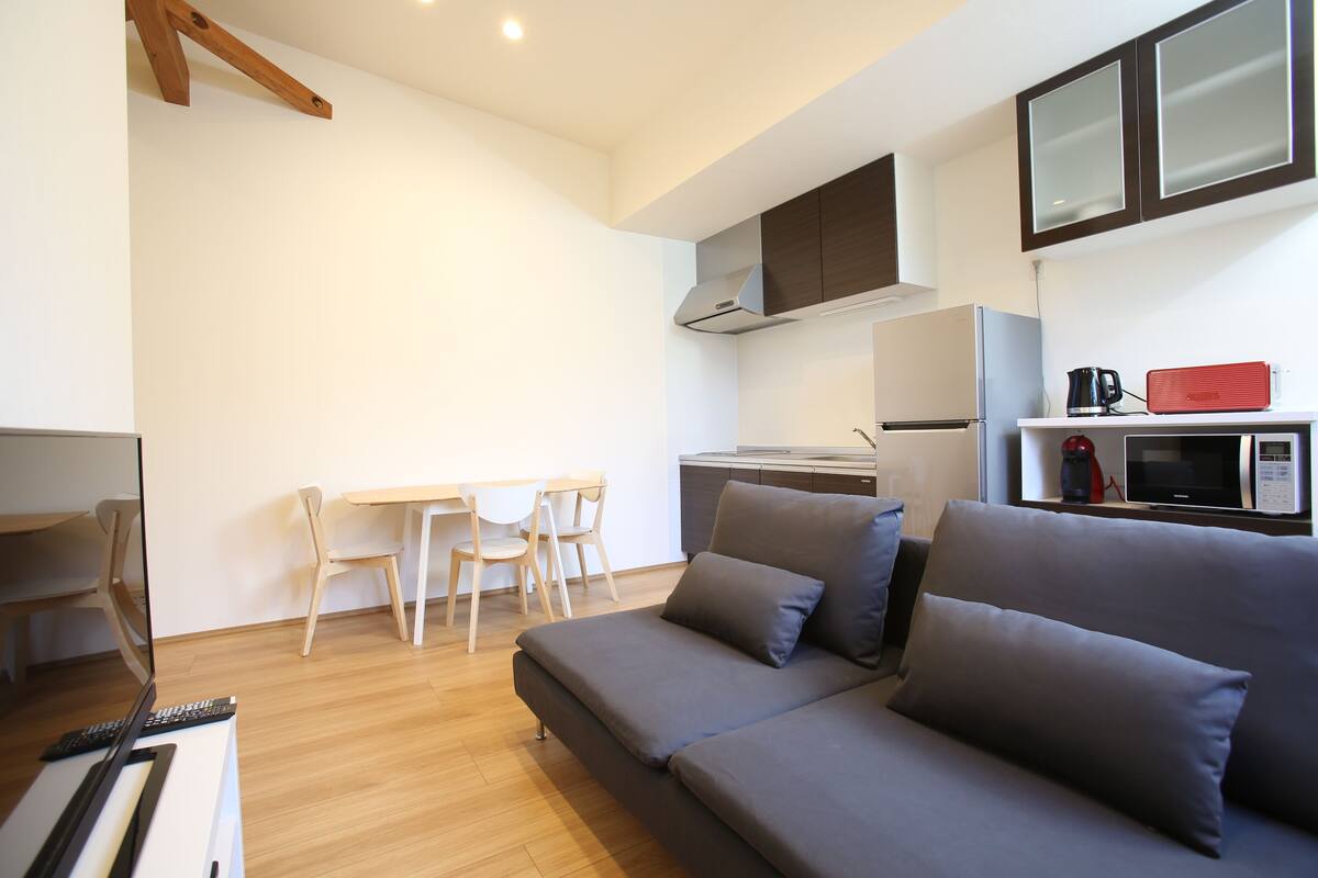 Uma sala de estar moderna com sofá, mesa de jantar com cadeiras e área de cozinha compacta com micro-ondas e cafeteira.