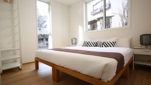 Ein Schlafzimmer mit einem hölzernen Bett, weißer Bettwäsche, einem Fenster mit Blick auf Gebäude, einem weißen Nachttisch und einer weißen Regaleinheit.