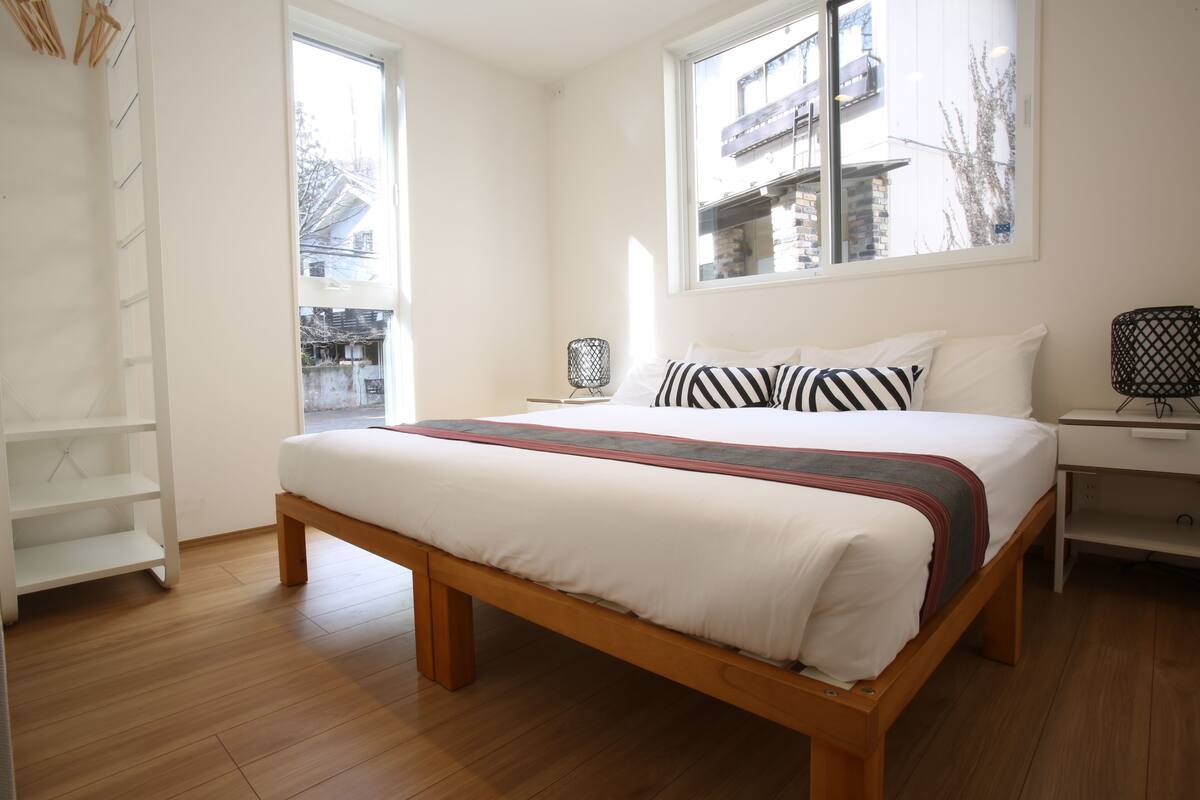 Quarto com cama de madeira, roupa de cama branca, janela com vista para prédios, mesa de cabeceira branca e estante branca.
