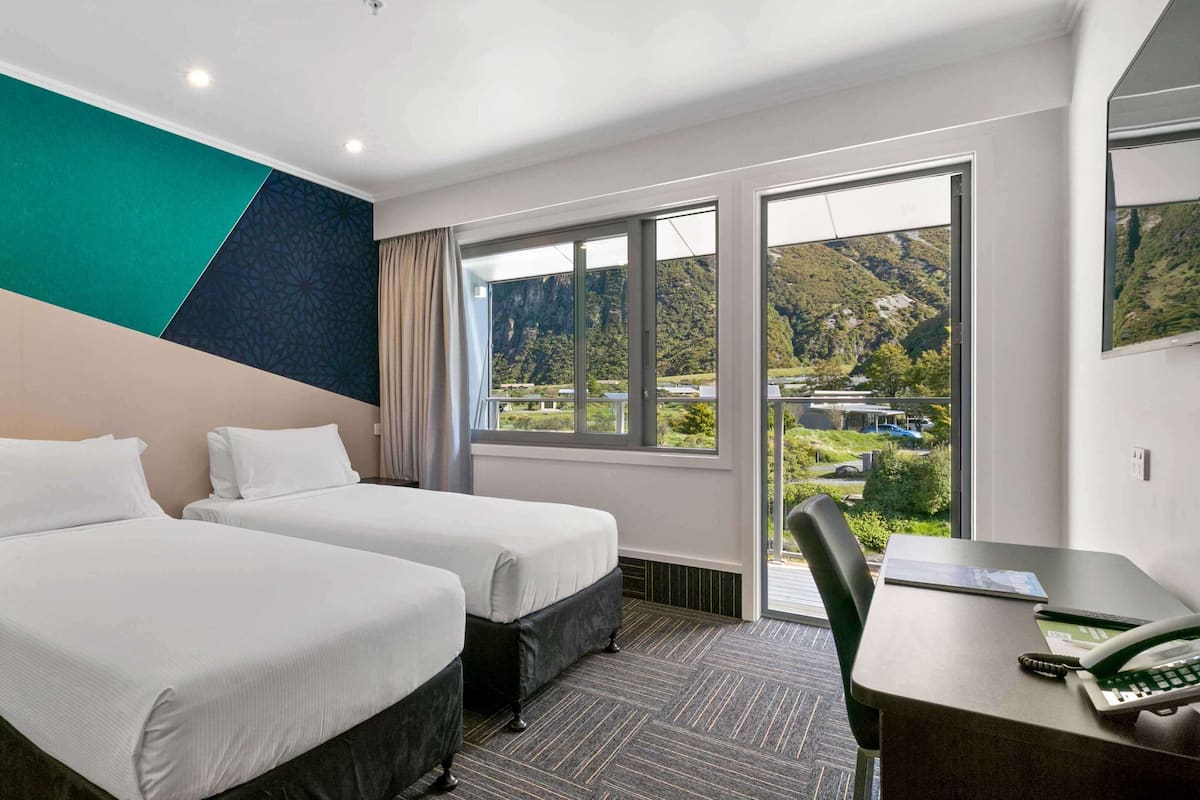 A hotel room with two beds, a desk, and a chair. There are large windows with a view of a mountainous landscape.