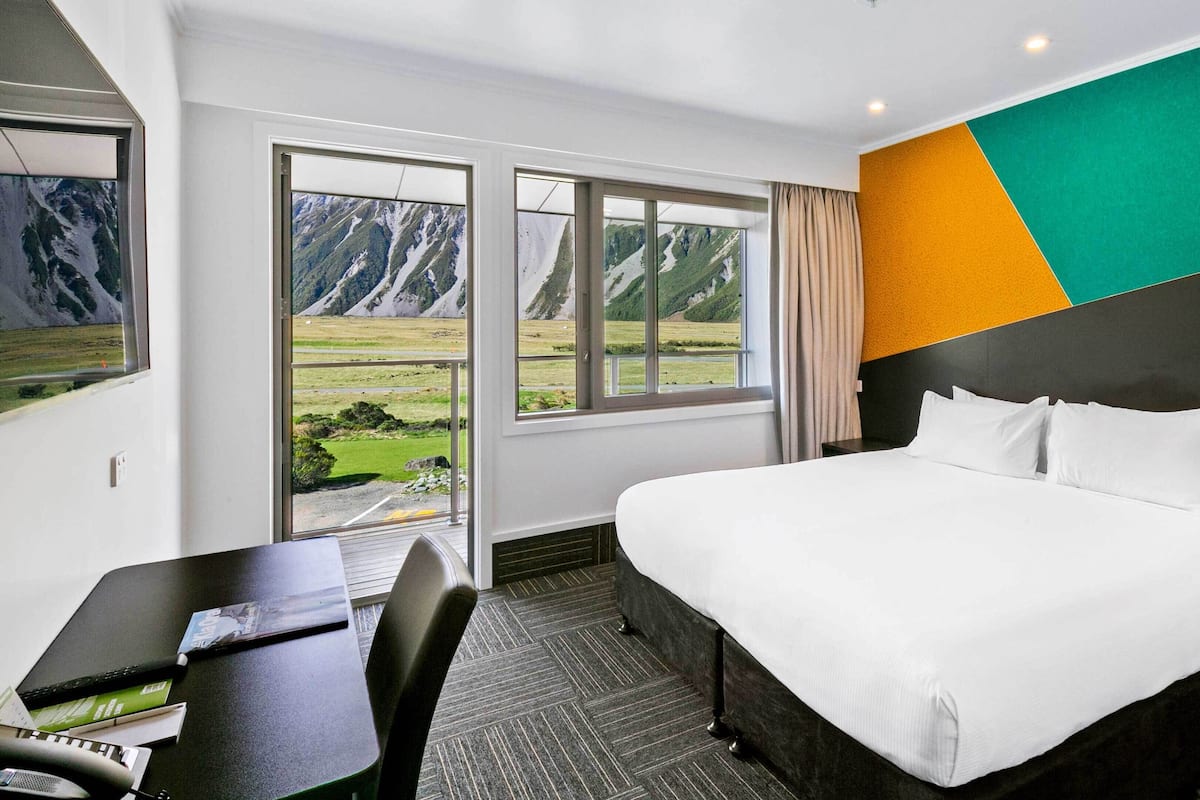 A hotel room with a large bed, a desk with a computer, and a view of mountains through the window.