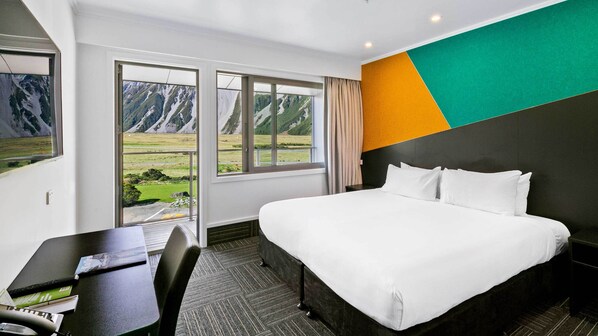 A hotel room with a large bed, a desk with a computer, and a view of mountains through the window. - Mt Cook Lodge & Motels (Mount Cook)