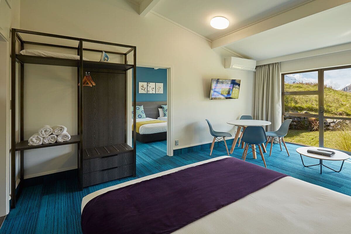 A hotel room with a bunk bed, a desk with chairs, a small table, and a large window with a view of the outdoors.
