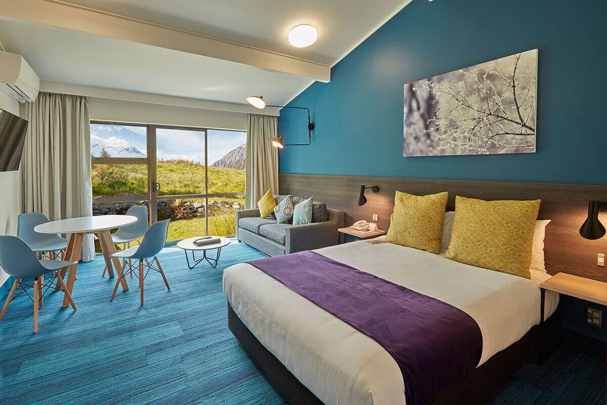 A hotel room with a large bed, a sofa, a dining table with chairs, and a view of mountains through large windows.