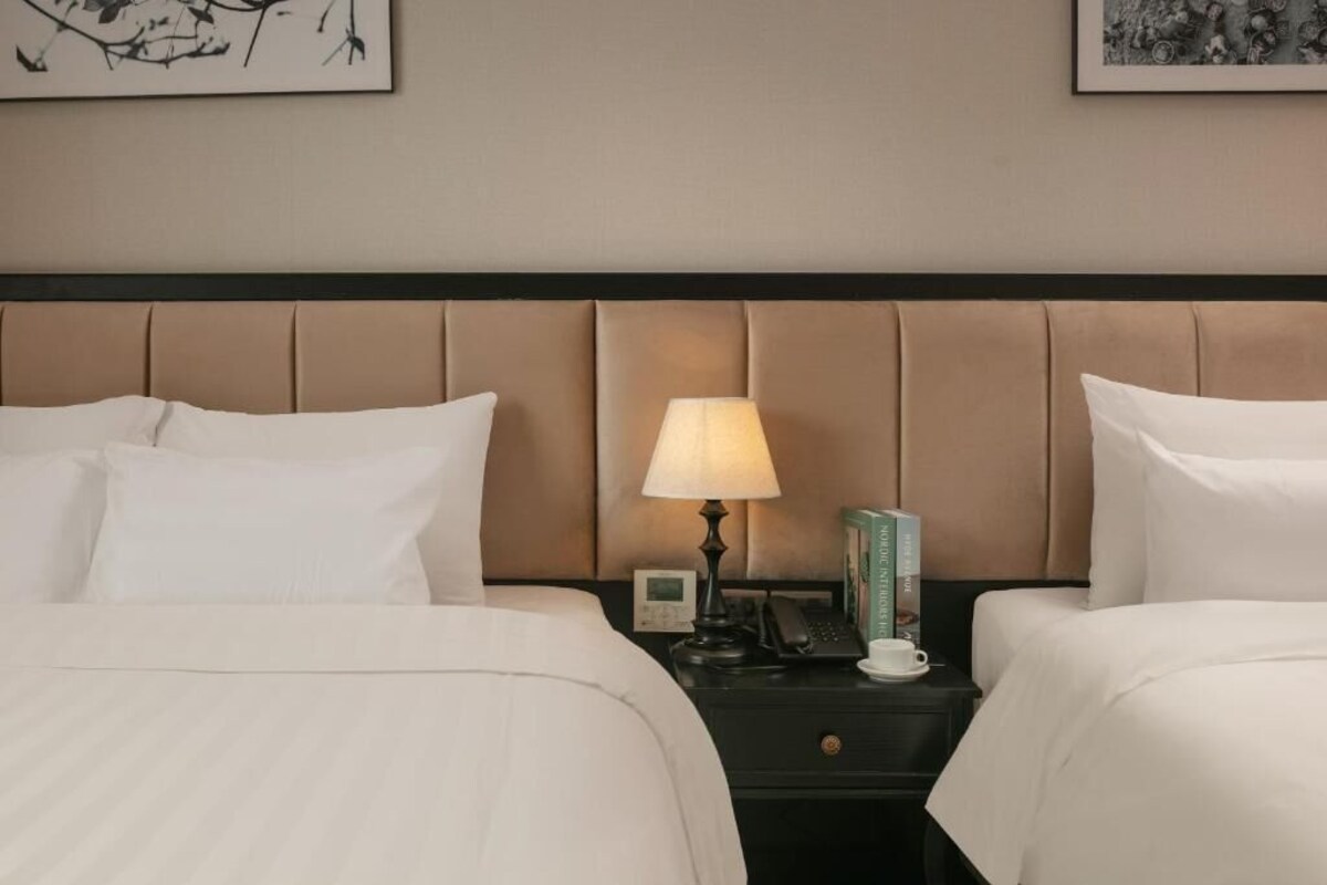 A hotel room with two beds, a nightstand with a lamp, a phone, a cup, and a picture frame.