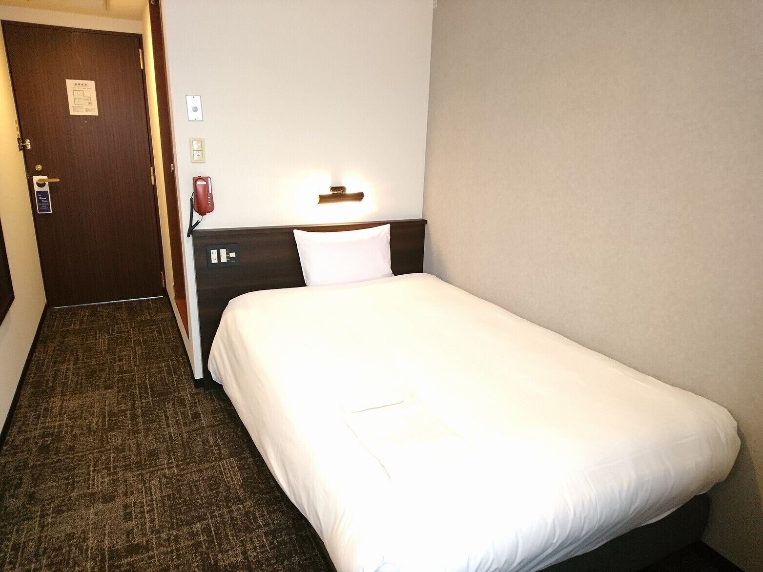 Ein Hotelzimmer mit einem Einzelbett, einem Nachttisch, einer Lampe, einem Telefon und einer Tür mit einem Schild.