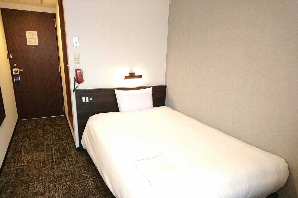 A hotel room with a single bed, a nightstand, a lamp, a telephone, and a door with a sign.