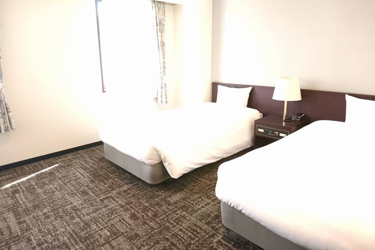 A hotel room with two beds, a nightstand, a lamp, and a wall-mounted clock.