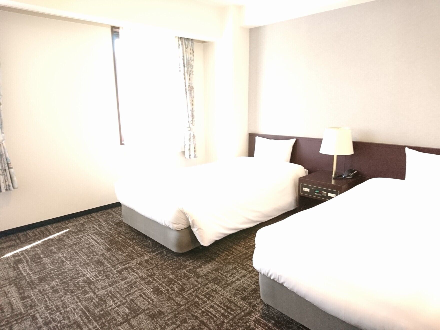 Ein Hotelzimmer mit zwei Betten, einem Nachttisch mit Lampe und einem Fenster mit Vorhängen.