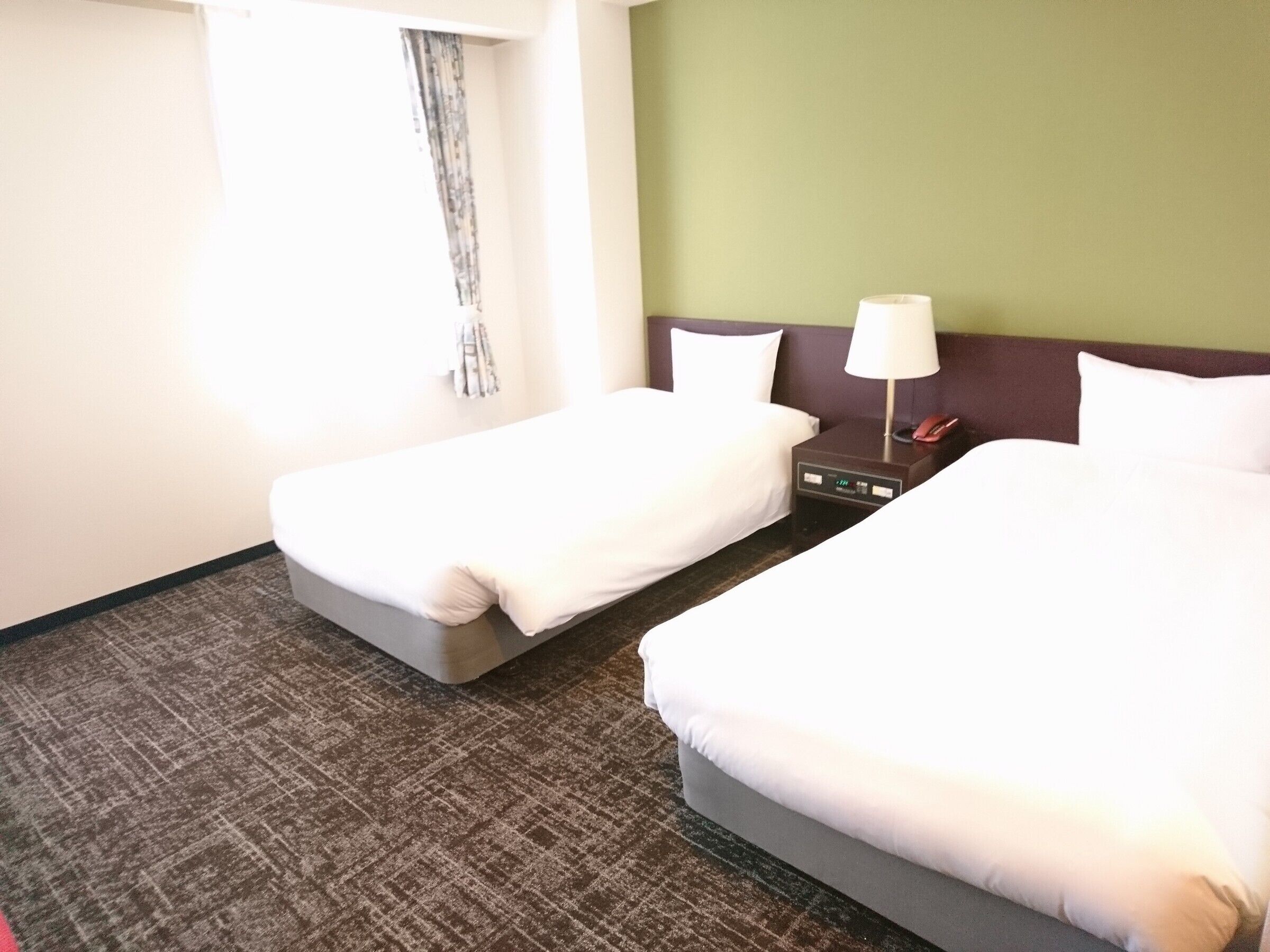 Ein Hotelzimmer mit zwei Betten, einem Nachttisch mit Lampe, einer Uhr und einem Fenster mit Vorhängen.
