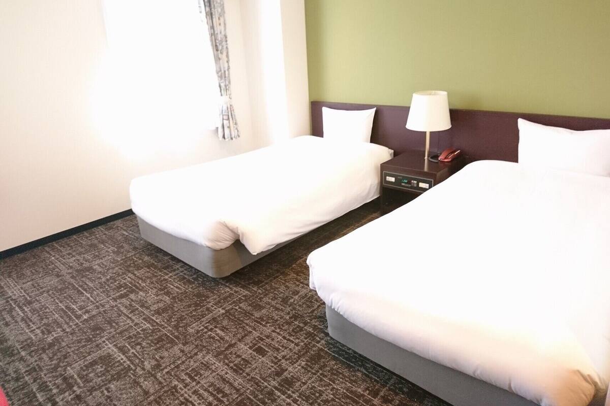 A hotel room with two beds, a nightstand with a lamp, and a window with curtains.