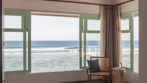 Ein Schlafzimmer mit einem großen Bett, einem Sessel, einem Nachttisch aus Holz und Meerblick durch Fenster mit grünen Rahmen.