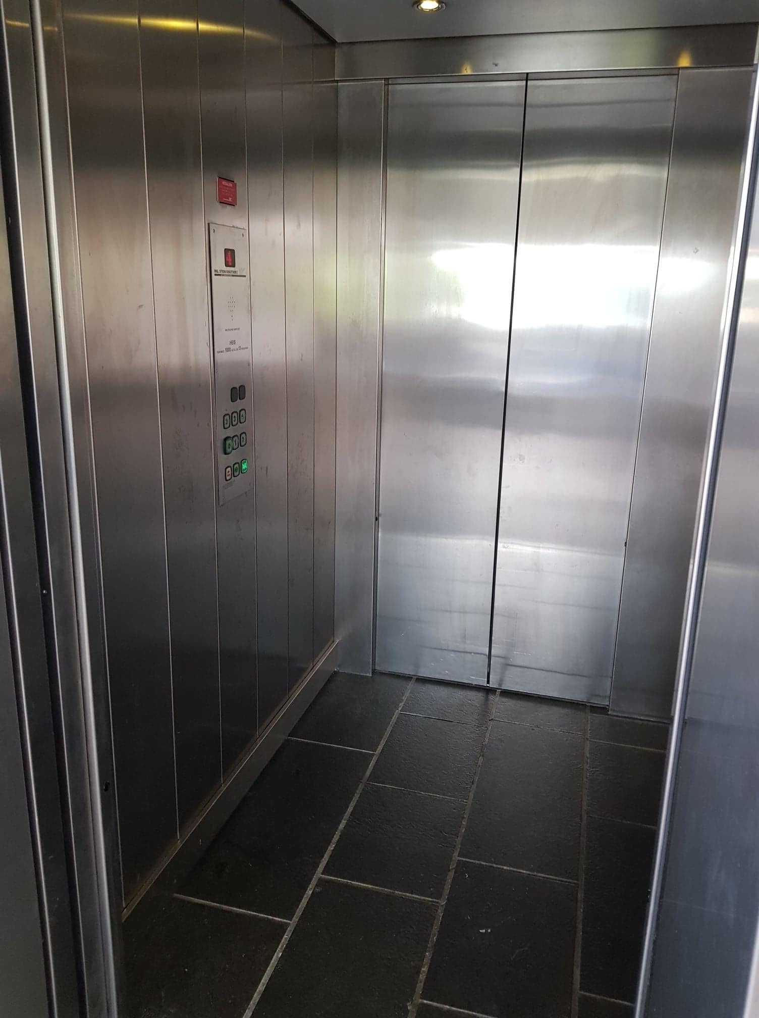 Interno di un ascensore con pareti metalliche, un pannello di controllo con pulsanti e pavimento piastrellato.