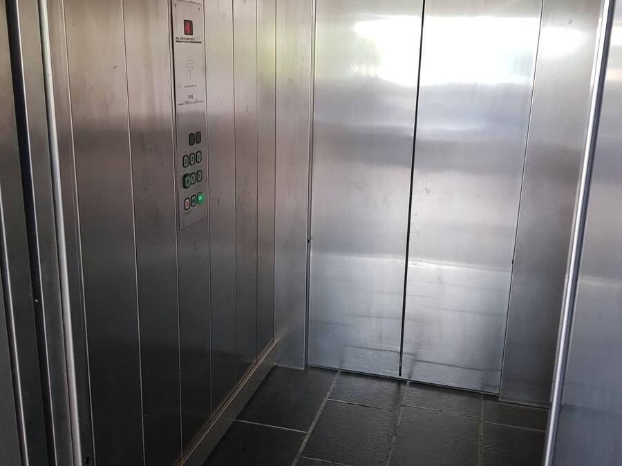 Interior de un ascensor con paredes metálicas, un panel de control con botones y suelo de baldosas.