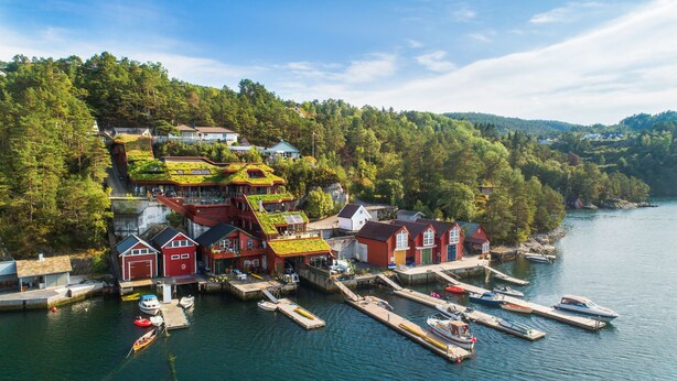 Uma vila costeira com casas tradicionais vermelhas, um cais com barcos e uma encosta arborizada.
