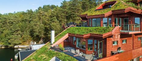 A multi-level wooden building with green roofs, large windows, and a view of a forested area and water body.