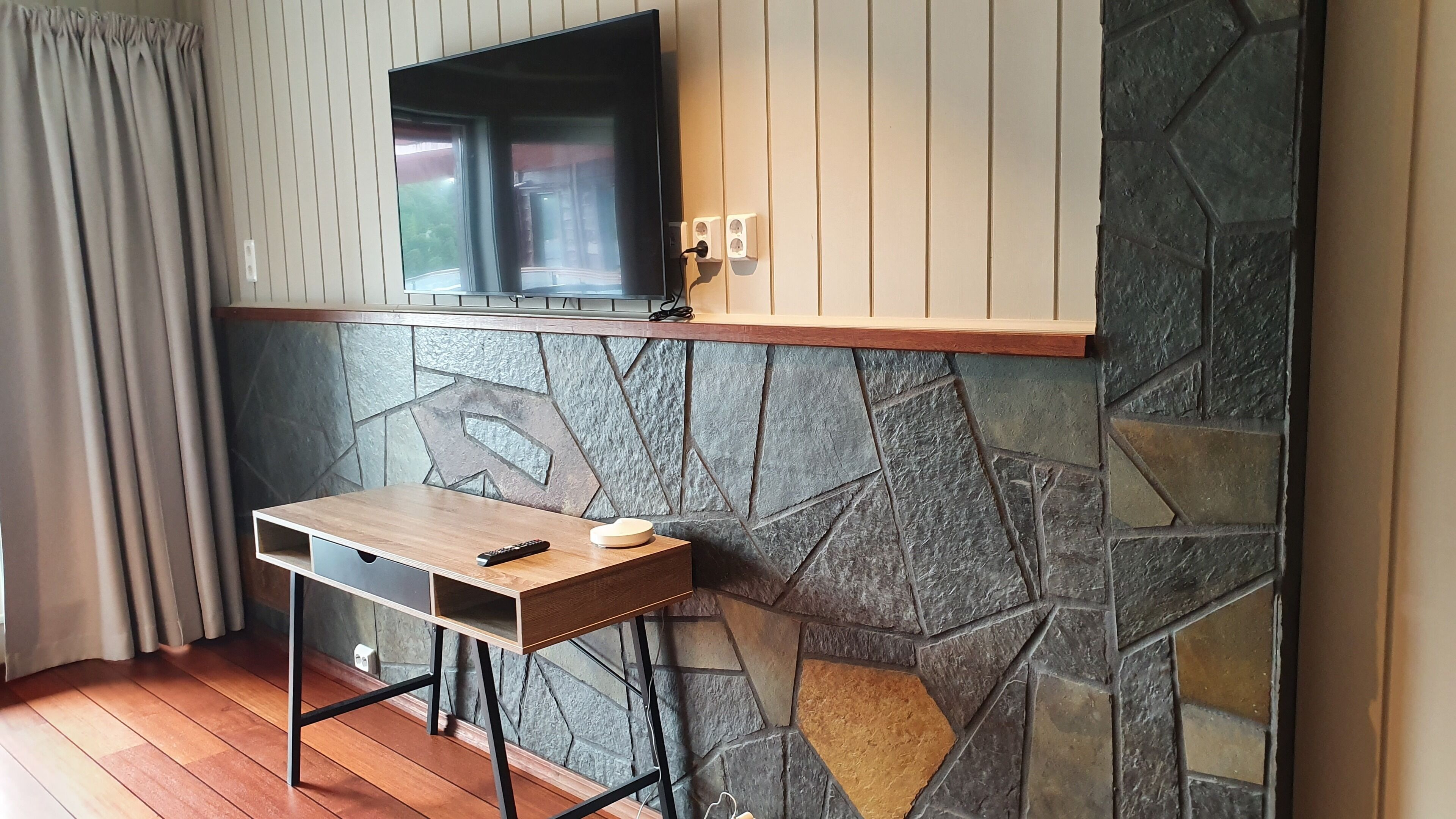 A room with a stone wall, a wooden table, a TV mounted on the wall, and a window with curtains.