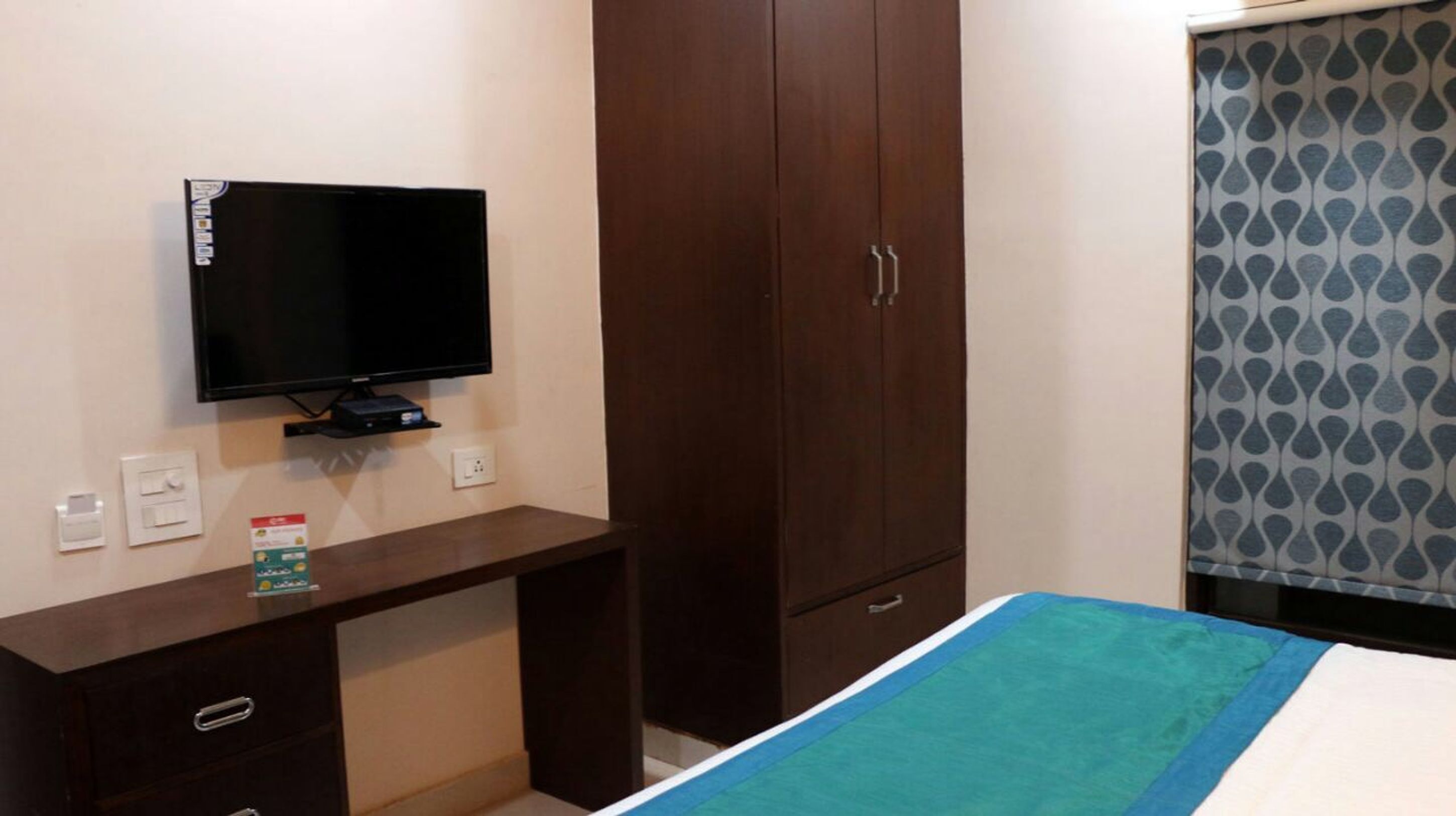 A bedroom with a bed, a TV on a stand, a wardrobe, and a desk with a bottle.