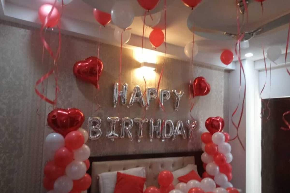 Een kamer met een bed, een muur versierd met 'HAPPY BIRTHDAY' ballonnen, en een rode ballon in de vorm van een hart.