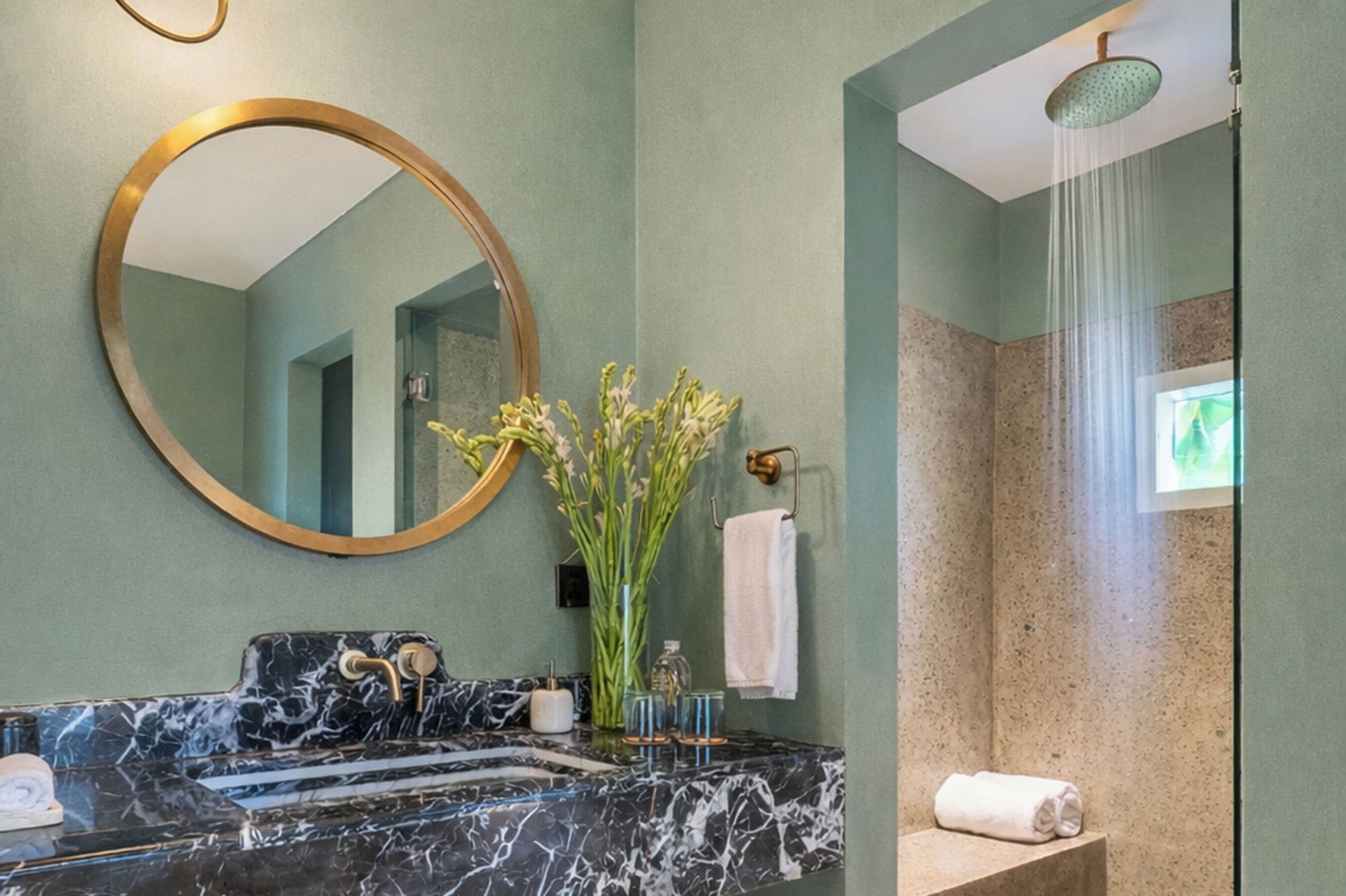 A bathroom with a marble sink, a large mirror, a shower, and a vase with flowers.