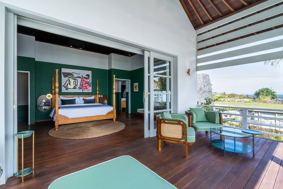 A bedroom with a bed, a green sofa, a glass table, and a view of the outdoors.